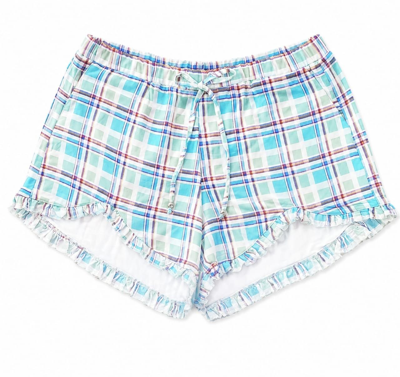 Mujer caminando por un sendero junto al mar con una blusa blanca y pantalones cortos de cuadros azules, calzando sandalias.