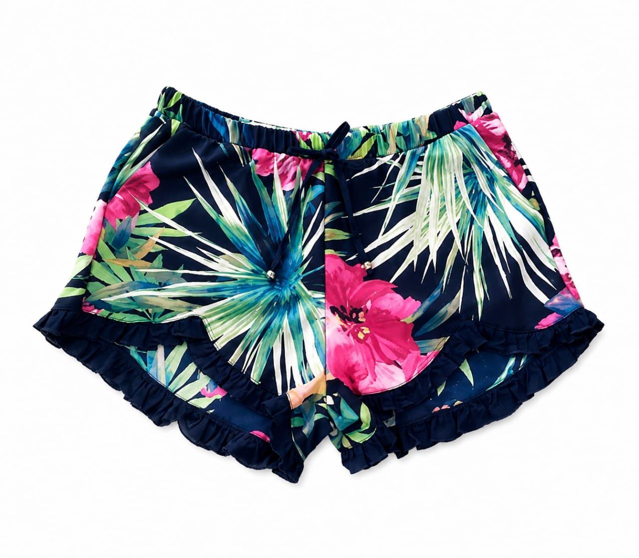 Mujer de pie en un sendero rocoso junto al agua, vestida con una blusa negra, pantalones cortos estampados con flores y sandalias azules.