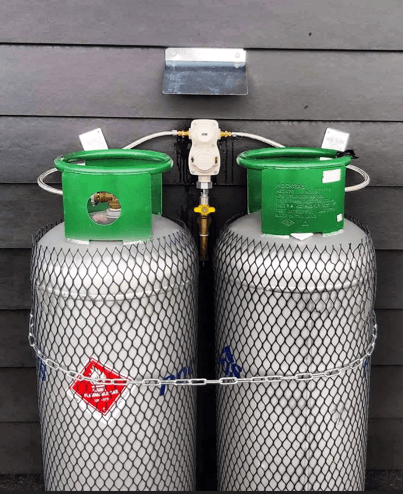 Two propane tanks connected to a regulator, mounted on a wall. A chain secures the tanks.