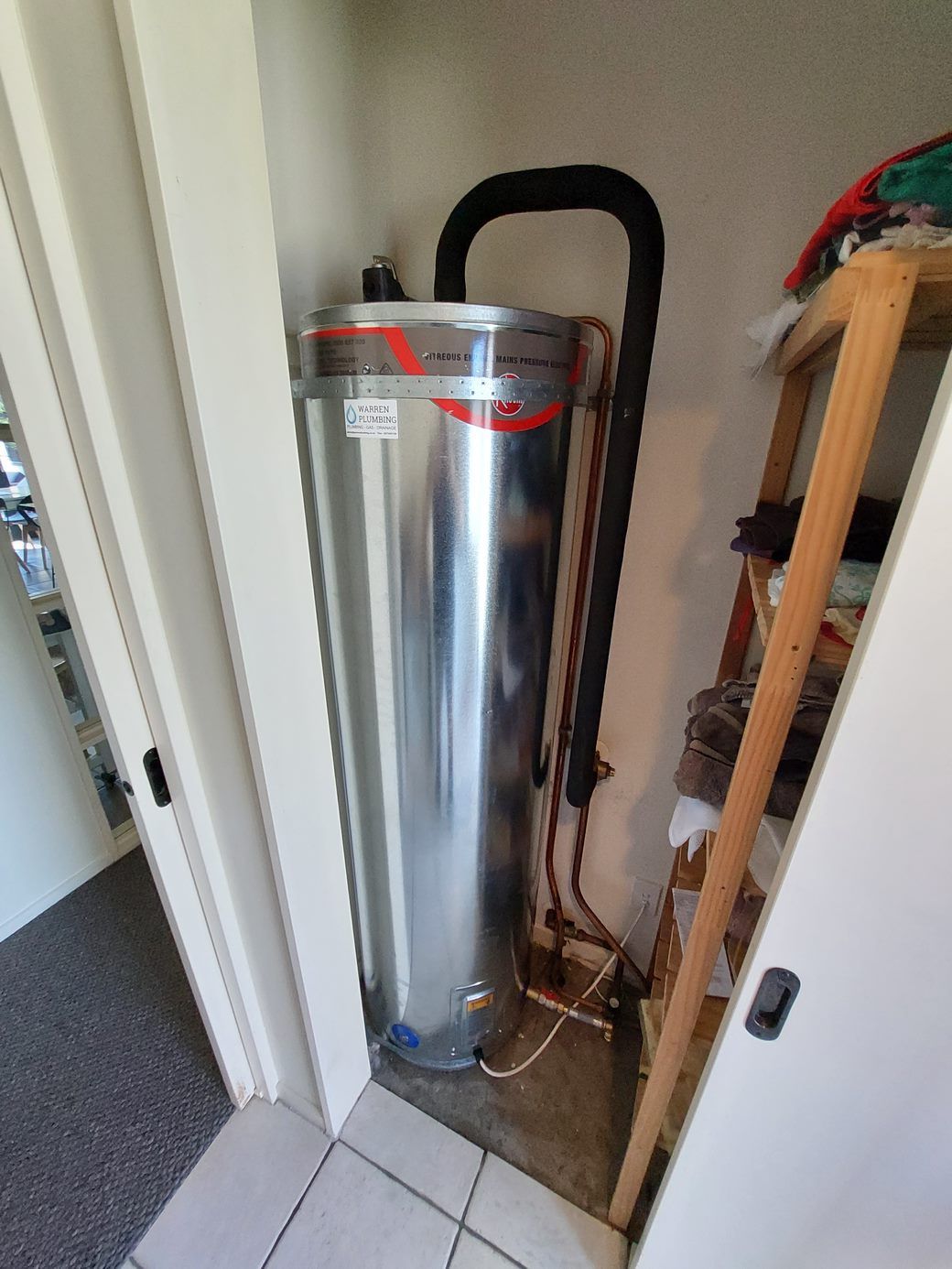 Tall, silver water heater in a small, narrow closet. A black pipe connects to the top, copper pipes below.
