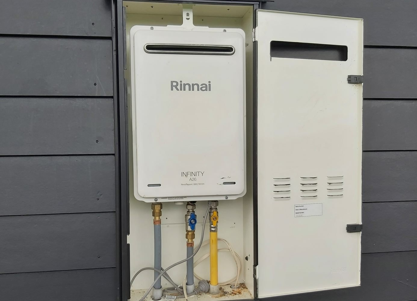 A white Rinnai tankless water heater mounted on a gray house exterior, with an open access panel revealing plumbing.
