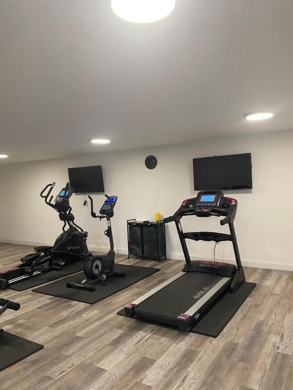 Gym with treadmill, elliptical, stationary bike, TVs, and equipment. Light wood-look floor, white walls.