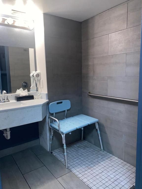 Accessible hotel bathroom with a shower, grab bar, and shower chair. Grey tile walls, white sink, blue chair.