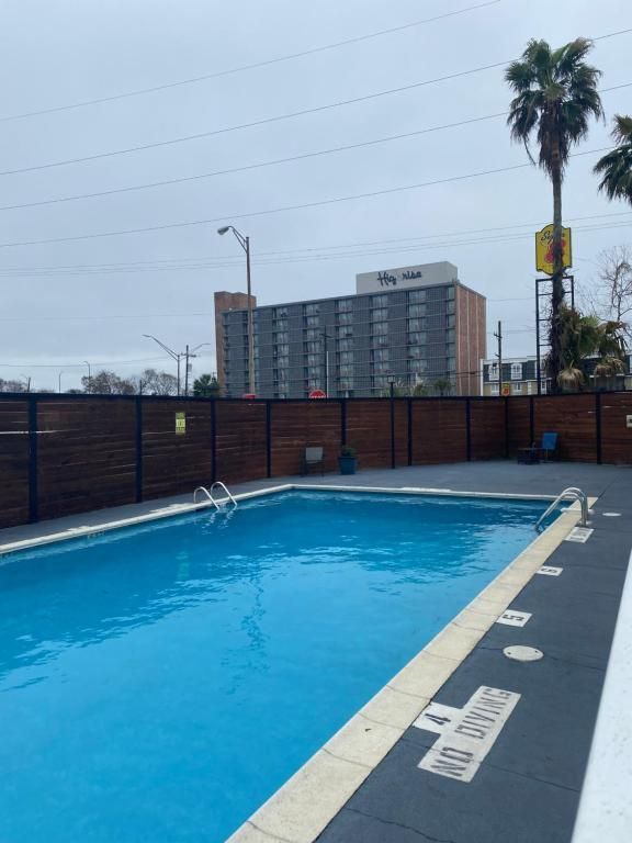 Outdoor swimming pool with blue water, surrounded by a wooden fence, with a tall building and palm trees in the background.
