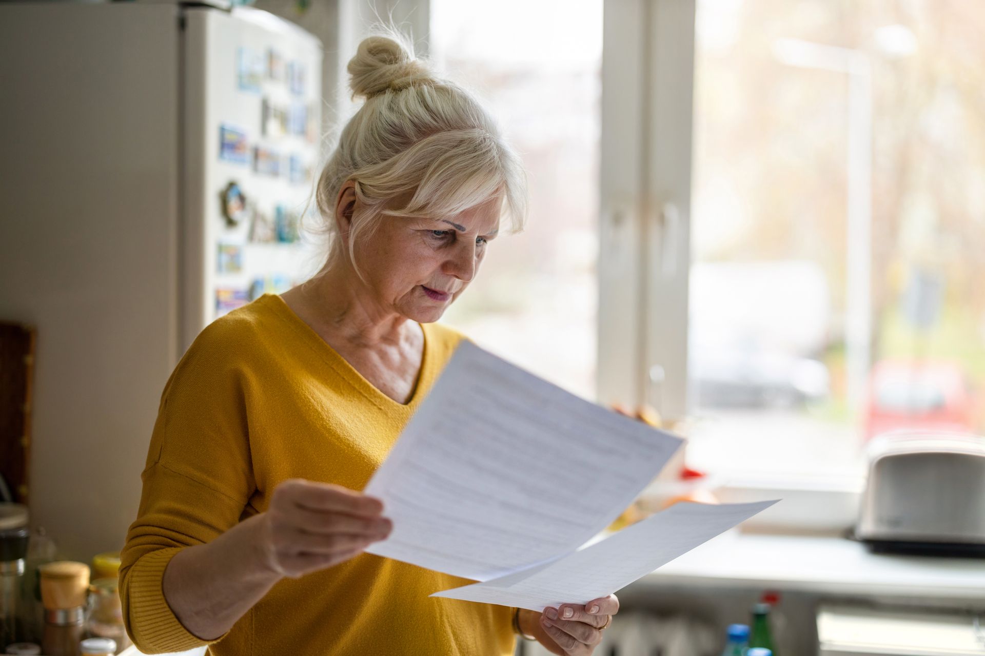 An elderly woman is reading a piece of paper in a kitchen.