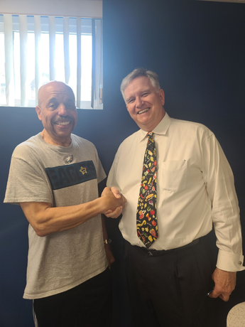 Two men smiling and shaking hands in an office, one in a gray T-shirt, one in a white shirt with a colorful tie.