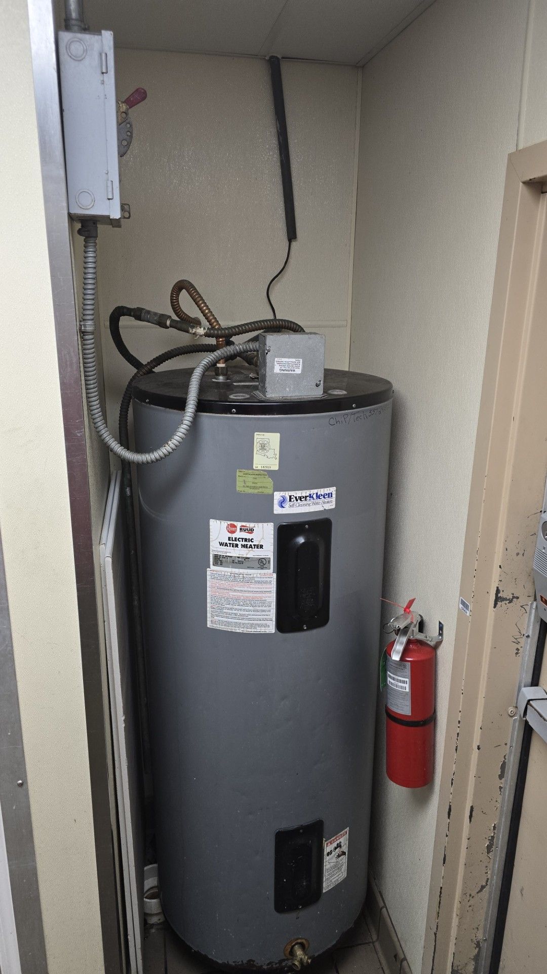 Water heater in a utility closet with exposed pipes and a red fire extinguisher on the wall.