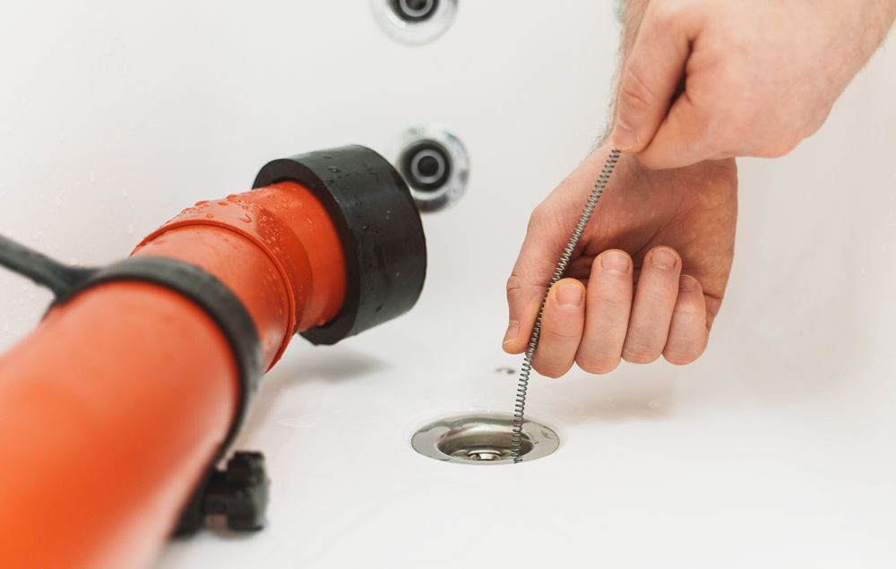 Hand unclogging a sink drain with a chain tool beside a red plunger