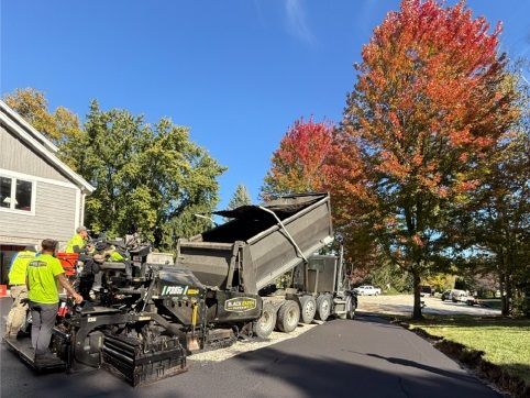 Two construction workers operating compact rollers on freshly paved asphalt near an industrial building.