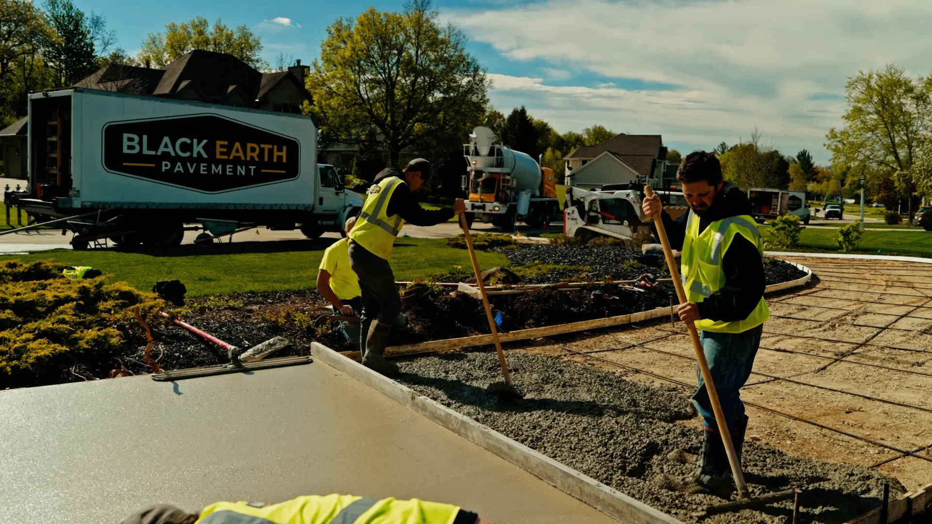 Workers in neon vests spread black earth at a roadside landscaping site beside a Black Earth truck.