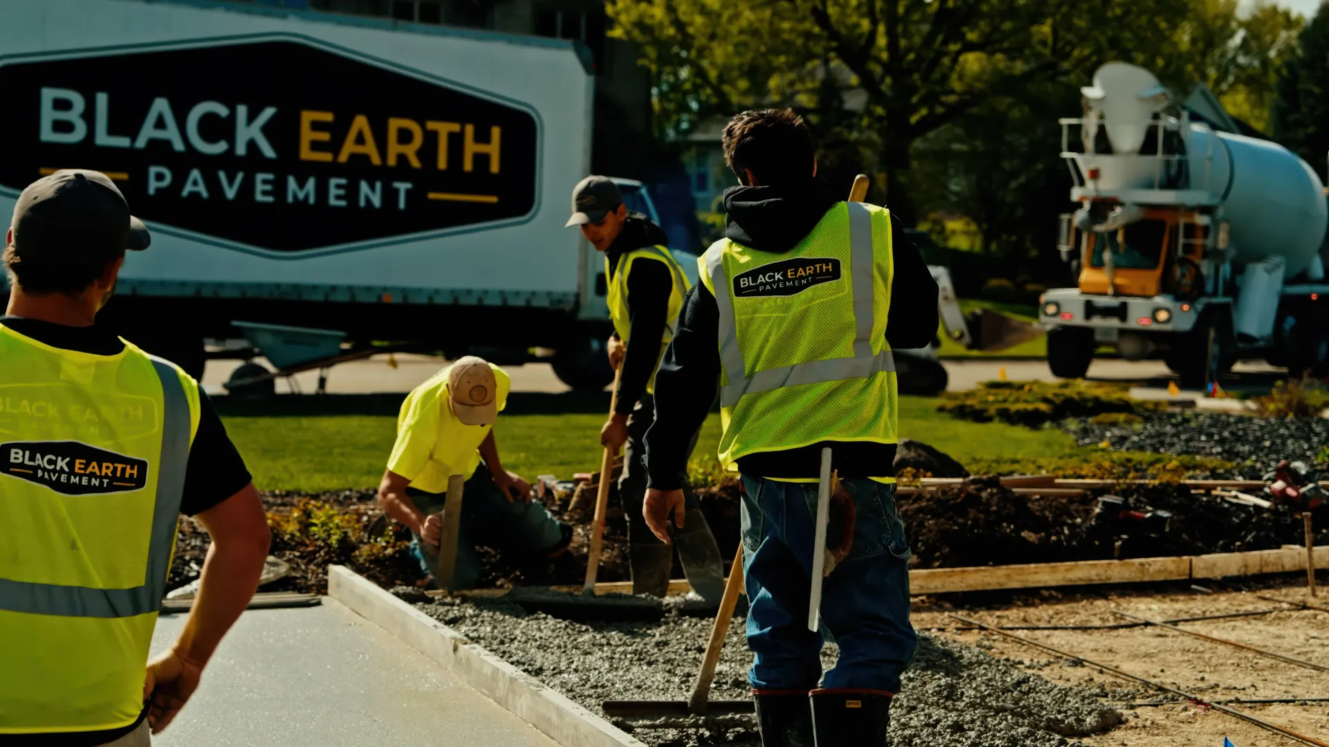 Two construction workers operating compact rollers on freshly paved asphalt near an industrial building.