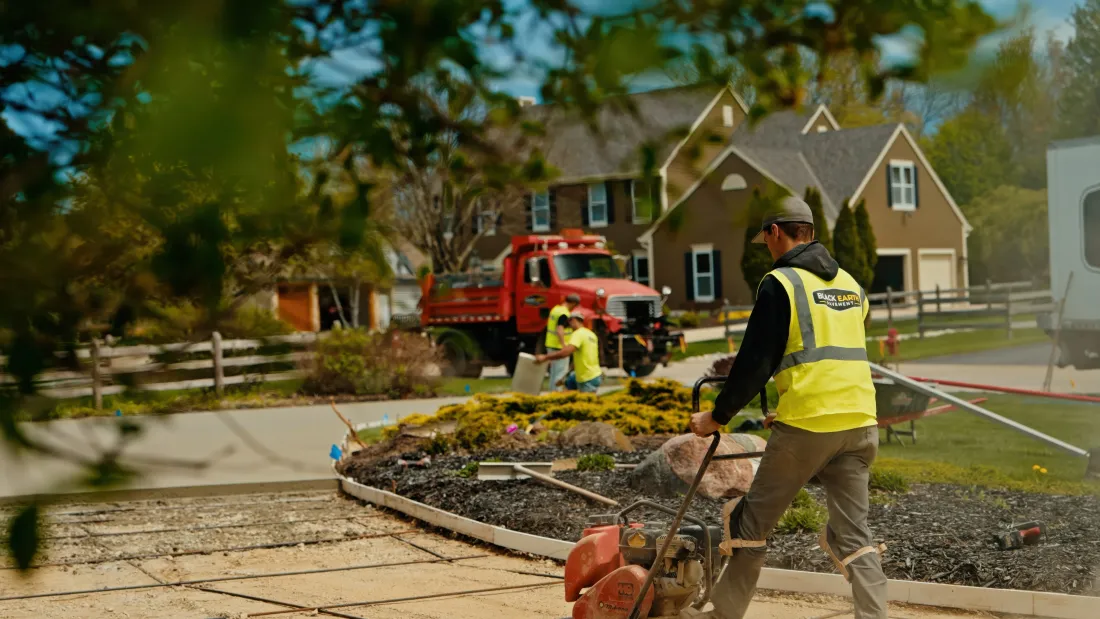 Worker in yellow vest pulls a wheelbarrow beside a road as a red truck sprays water near suburban houses.