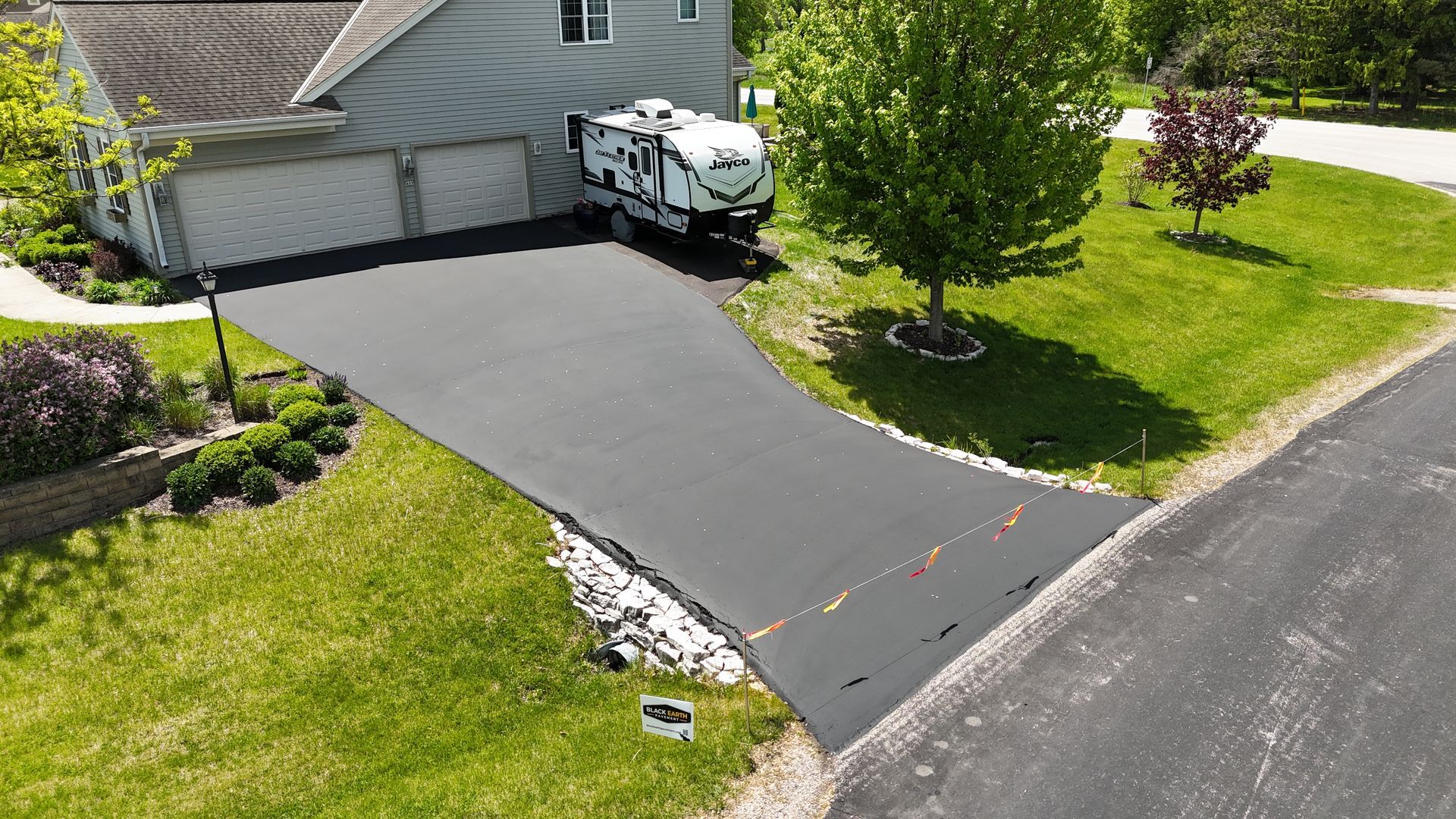 Freshly paved black driveway beside a gray house, with a parked boat and green lawn by the road