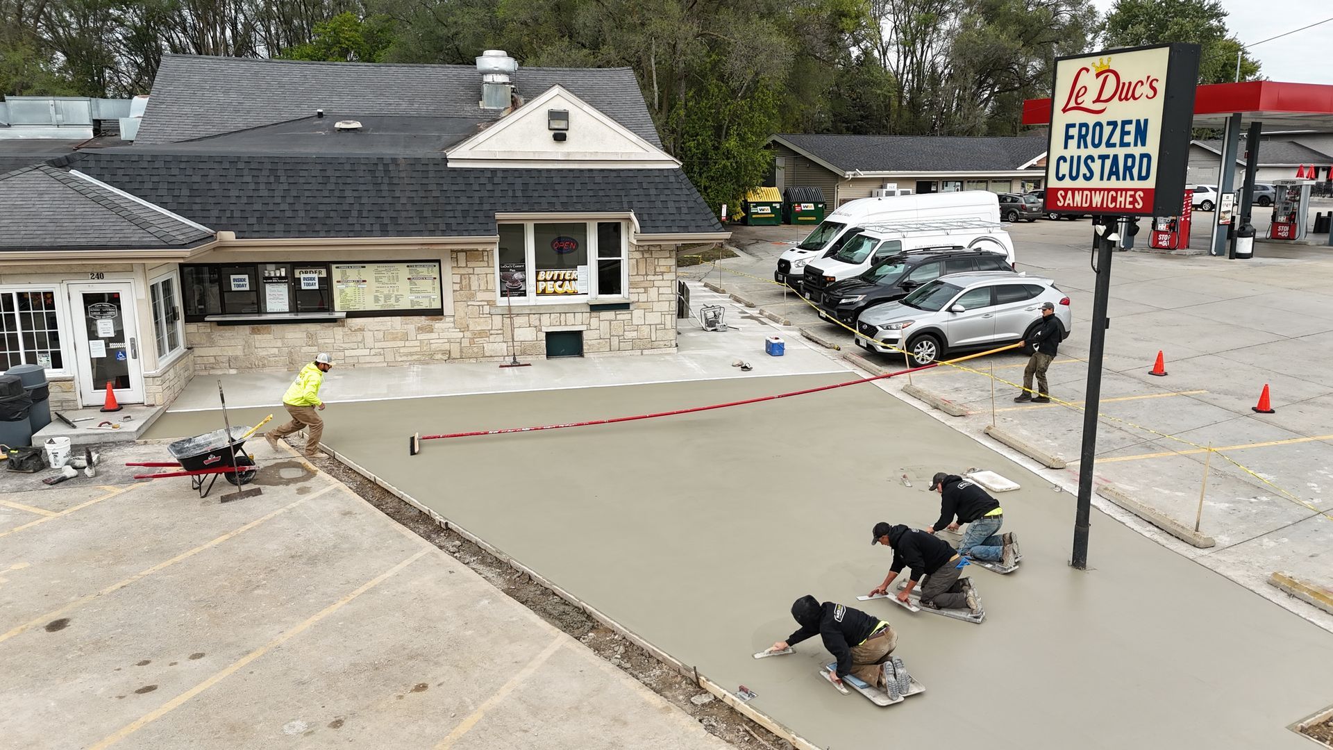 Workers repaving a parking lot beside a cafe with a large sign, cones, and parked cars