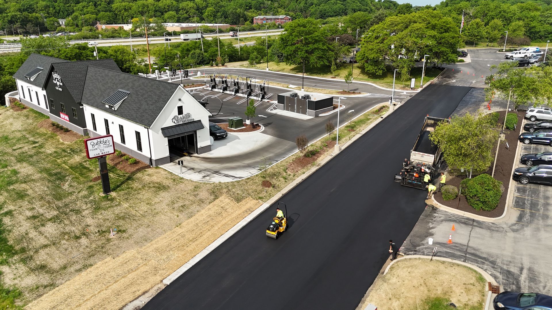 Aerial view of a road construction site with a black paved road, yellow machinery, and a white building nearby