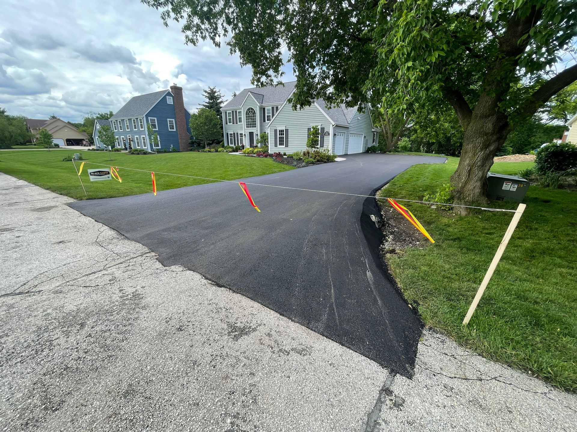 Freshly paved driveway curves toward a white house, with orange survey stakes and trees along the lawn.