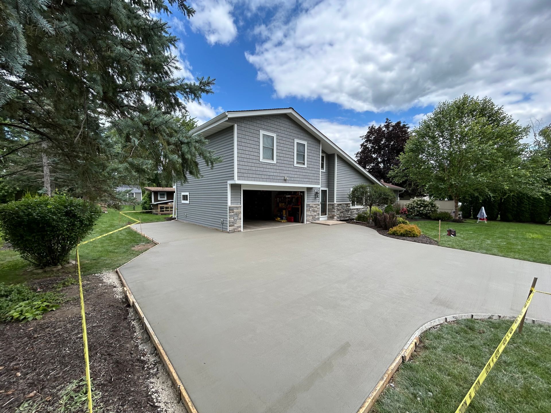 Gray house with open garage and wide concrete driveway, surrounded by green lawn and trees under cloudy sky