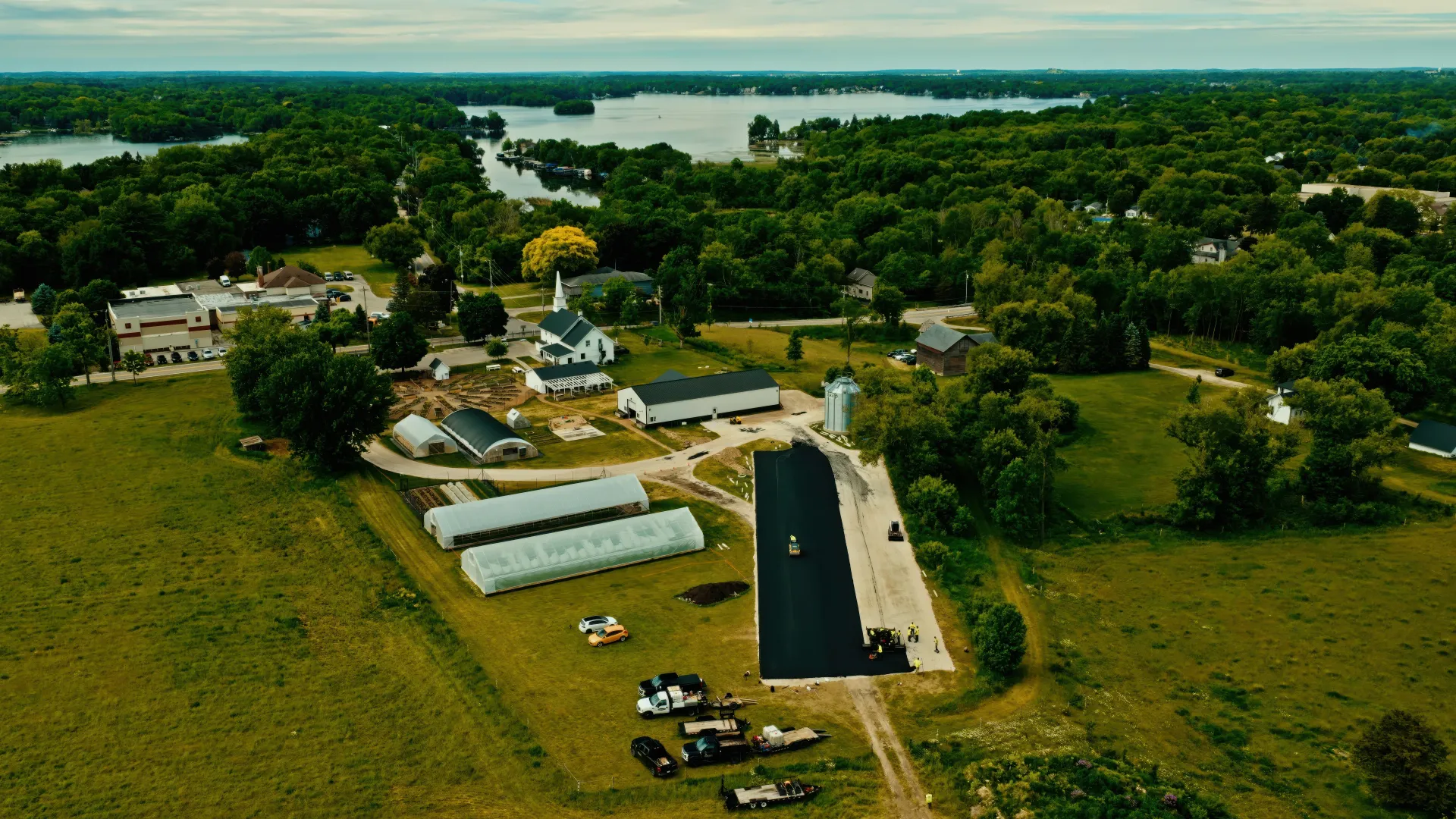 Aerial view of a rural farm with green fields, white buildings, and a long black-roofed structure near water.