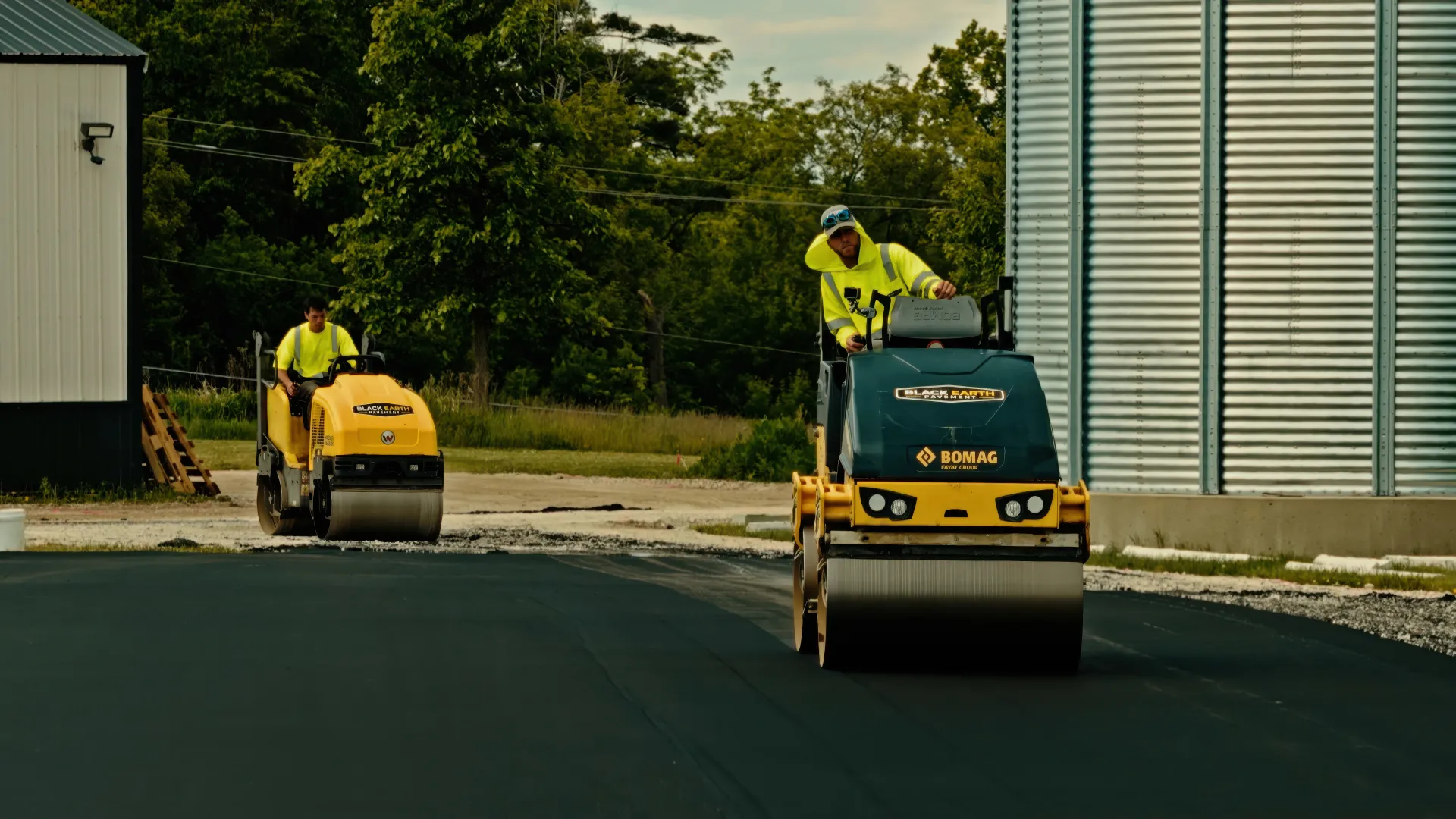 Two construction workers operating compact rollers on freshly paved asphalt near an industrial building.