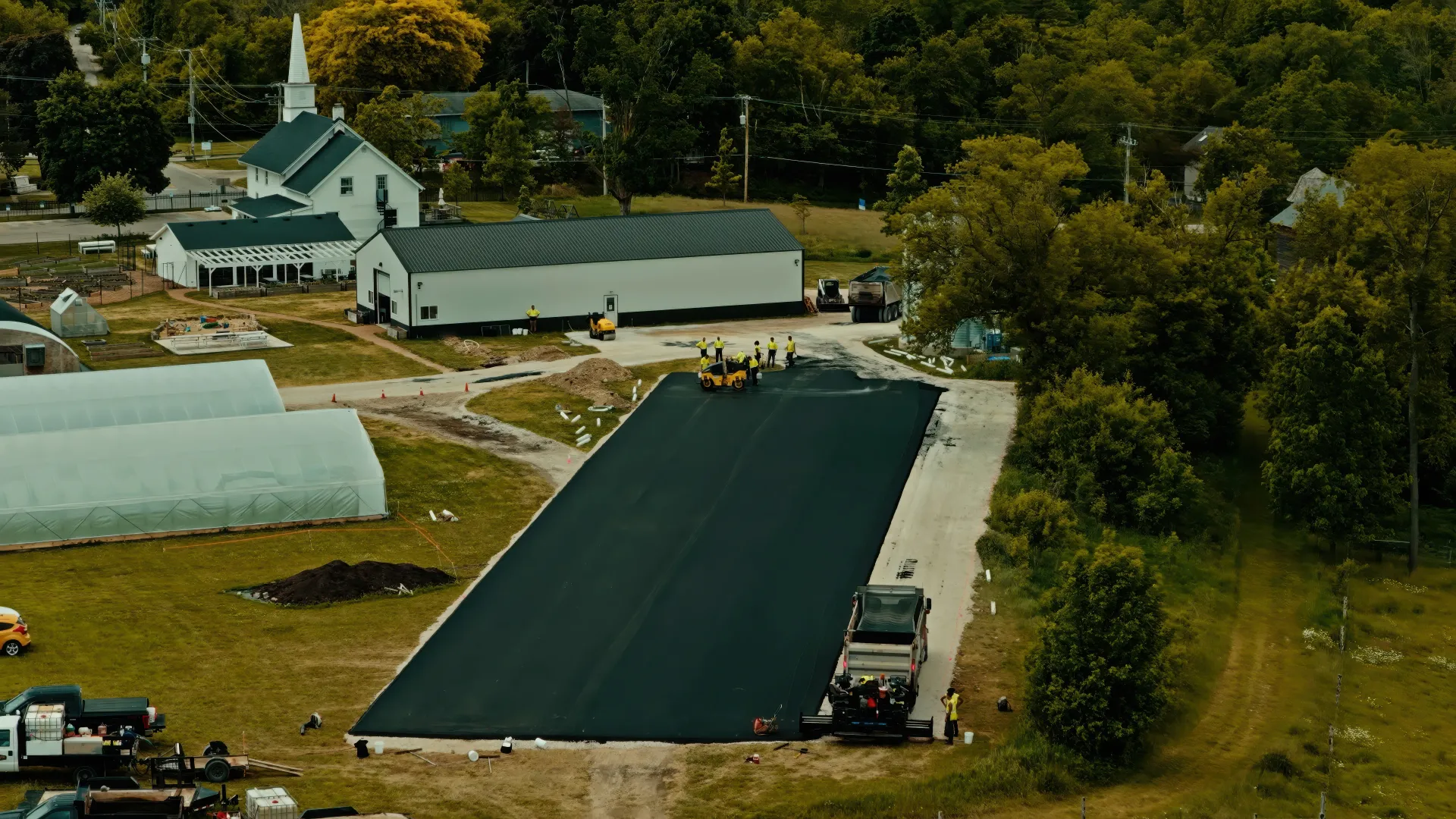 Aerial view of a farm with greenhouses, barns, and a long dark paved path surrounded by trees.