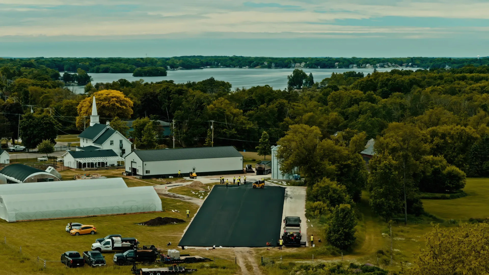 Wide aerial view of a rural farm with a white barn, dark-roofed buildings, trees, and a lake beyond