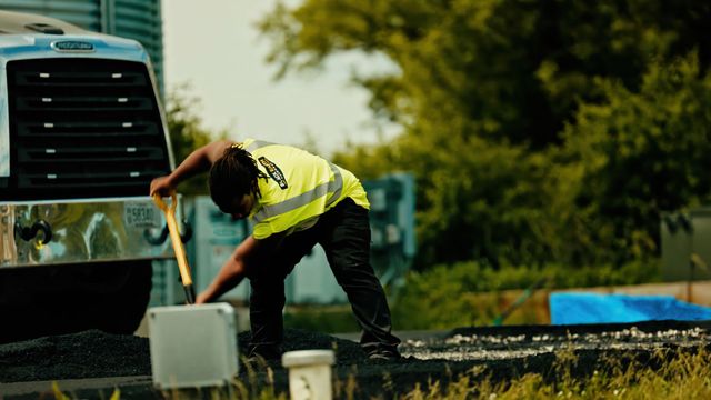Worker in a neon vest bends beside a truck, handling a white container outdoors.