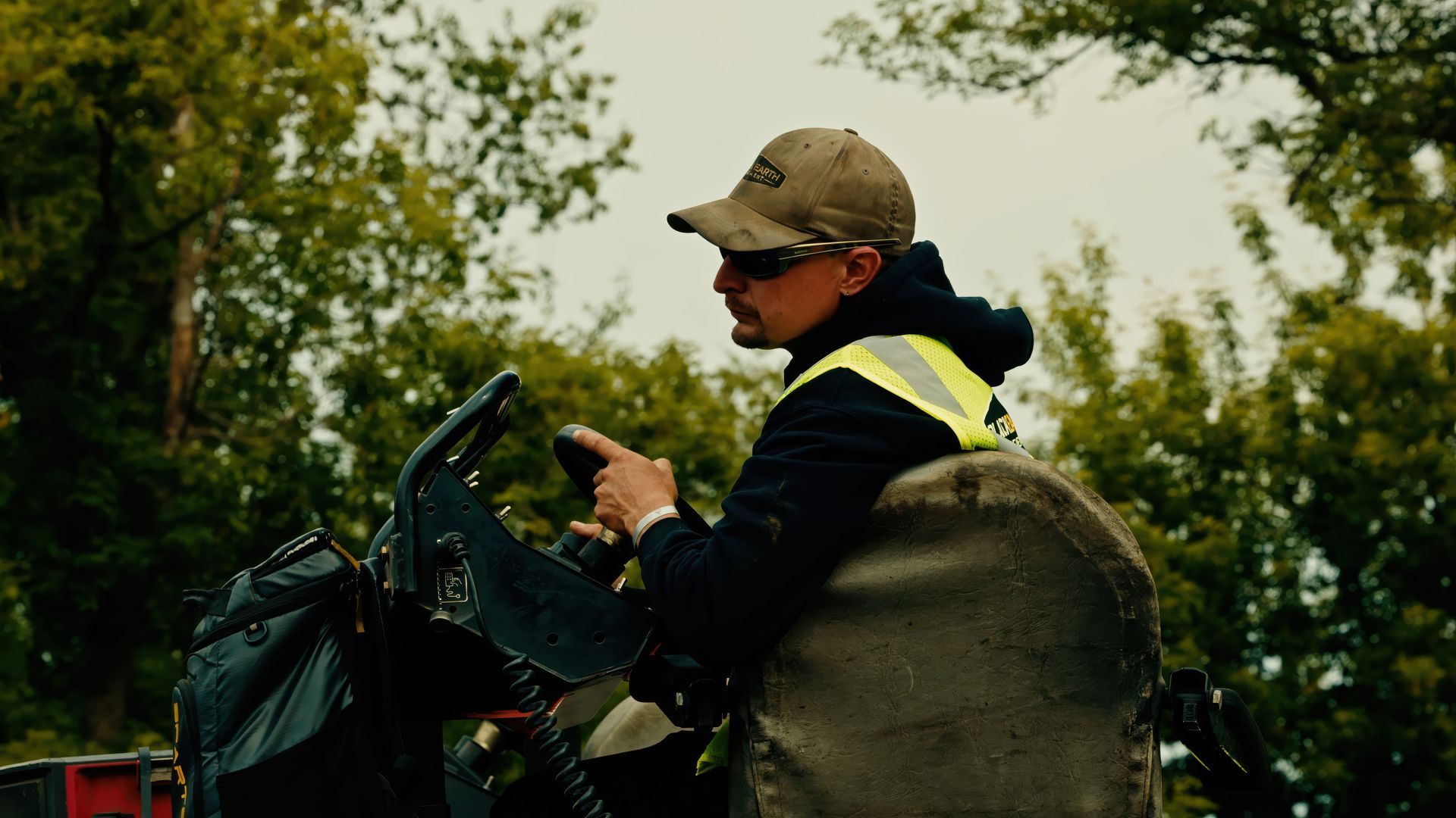 Person in a camo cap and safety vest drives an ATV through a wooded area.