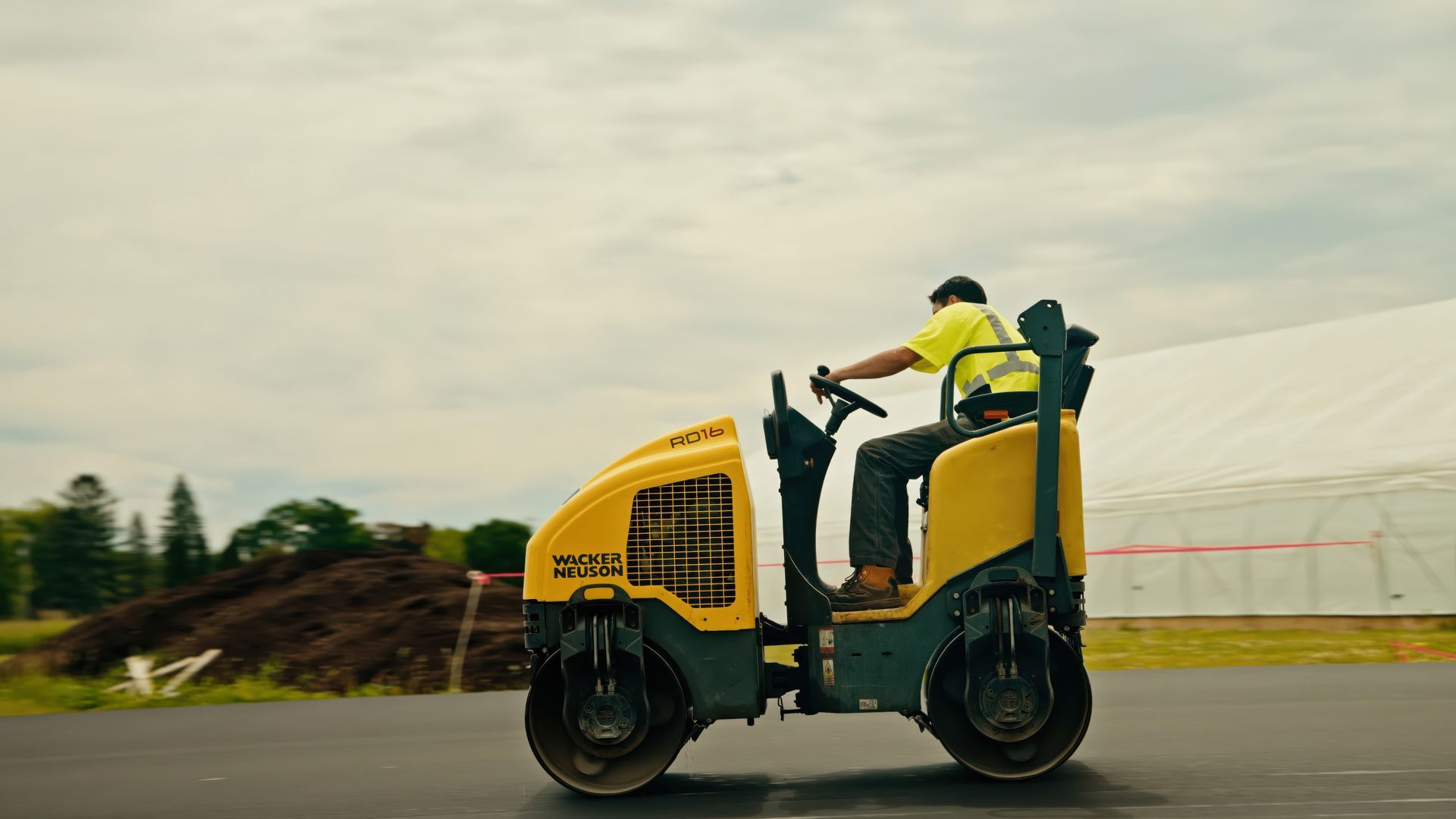 Yellow road roller with an operator driving on a paved site, with a cloudy sky and construction area in the background