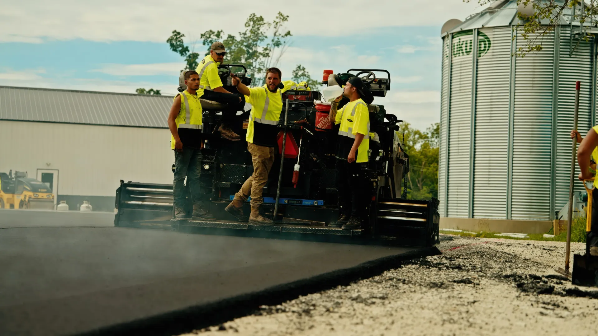 Workers in yellow shirts ride on a truck spraying dust along a dirt road beside grain silos.