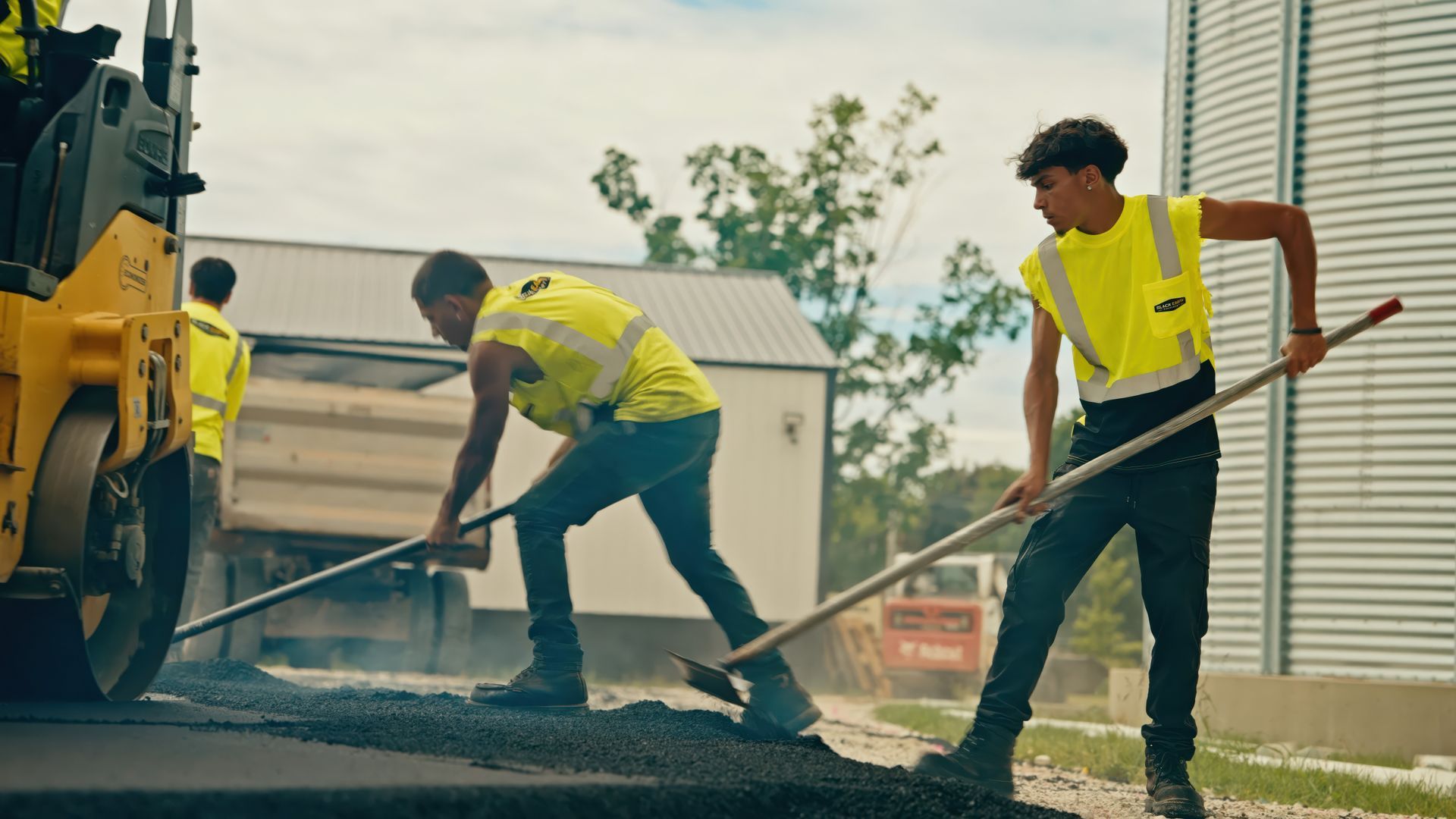 Two construction workers operating compact rollers on freshly paved asphalt near an industrial building.