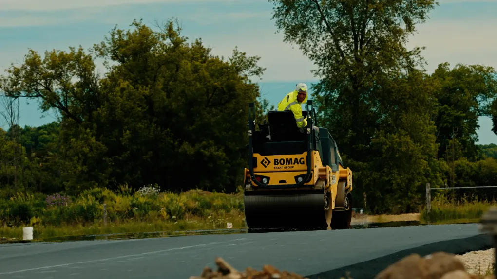 Yellow road roller on a rural road with a worker in a neon vest, trees in the background