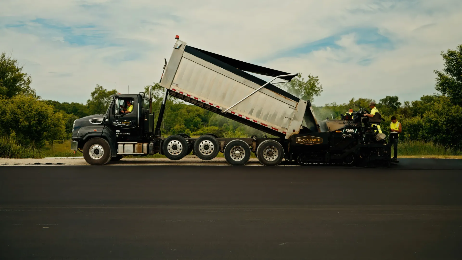 truck unloading gravel beside a paved road, with trees and cloudy sky in the background