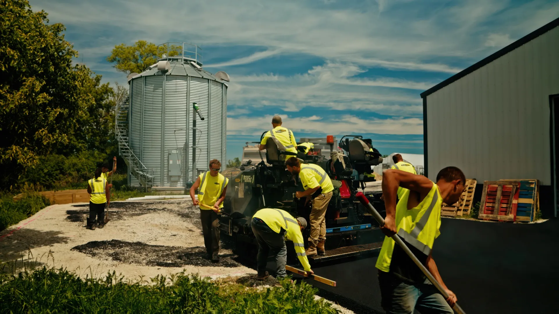 Workers in yellow safety gear load equipment beside a large metal silo and building outdoors.