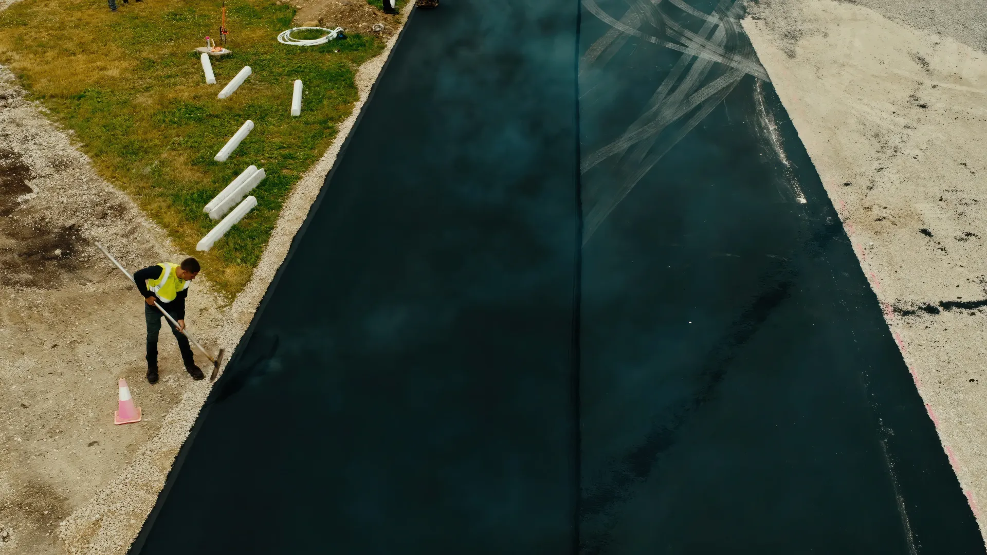 Aerial view of a worker painting a dark surface beside scattered white materials on a gravel edge.