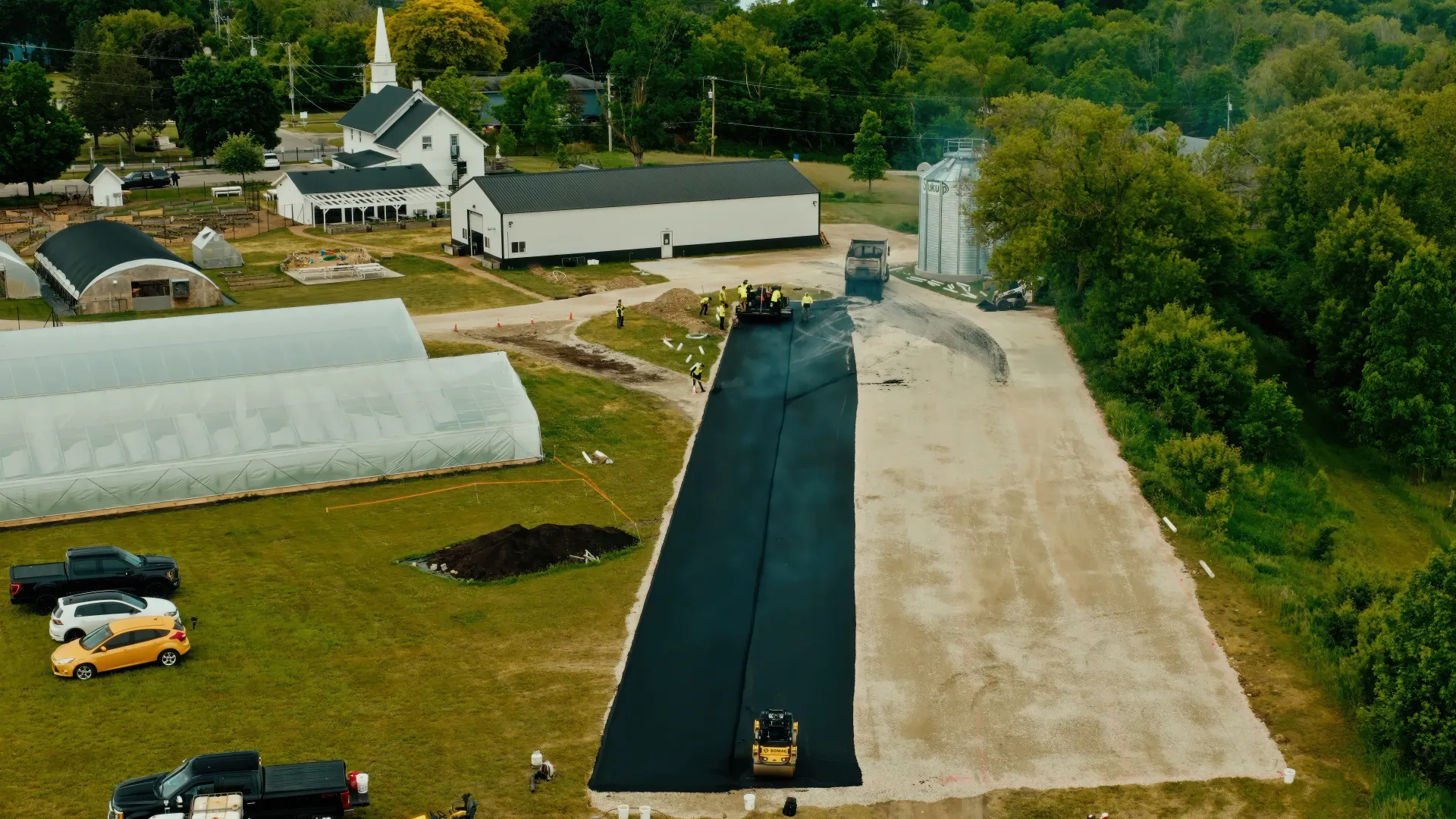 Aerial view of a farm with greenhouses, barns, and a long black paved strip under construction