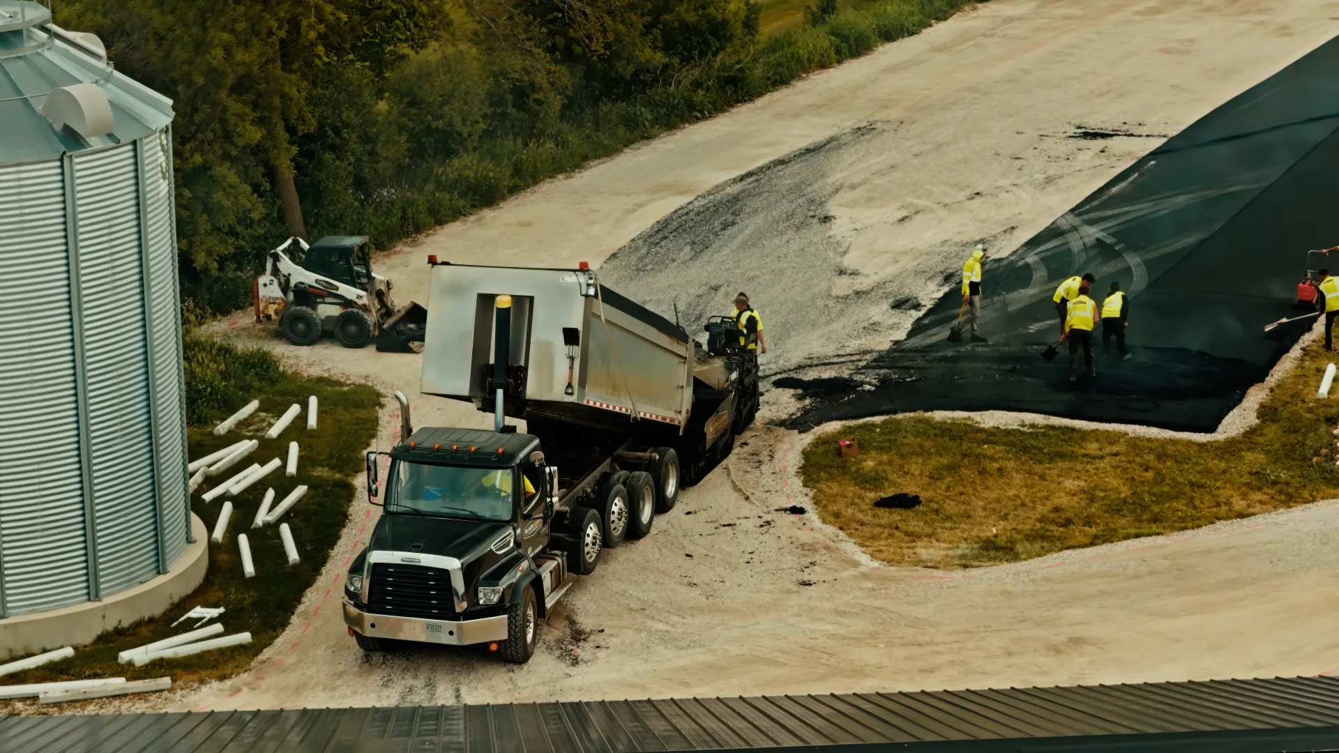 Two construction workers operating compact rollers on freshly paved asphalt near an industrial building.