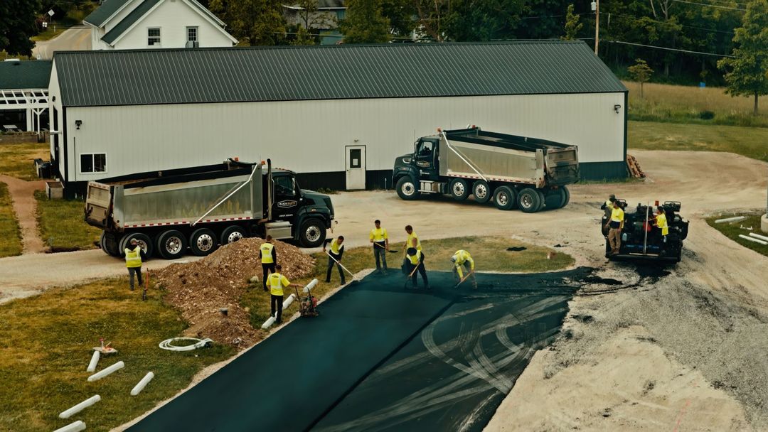 Workers and trucks at a roadside construction site beside a large white building