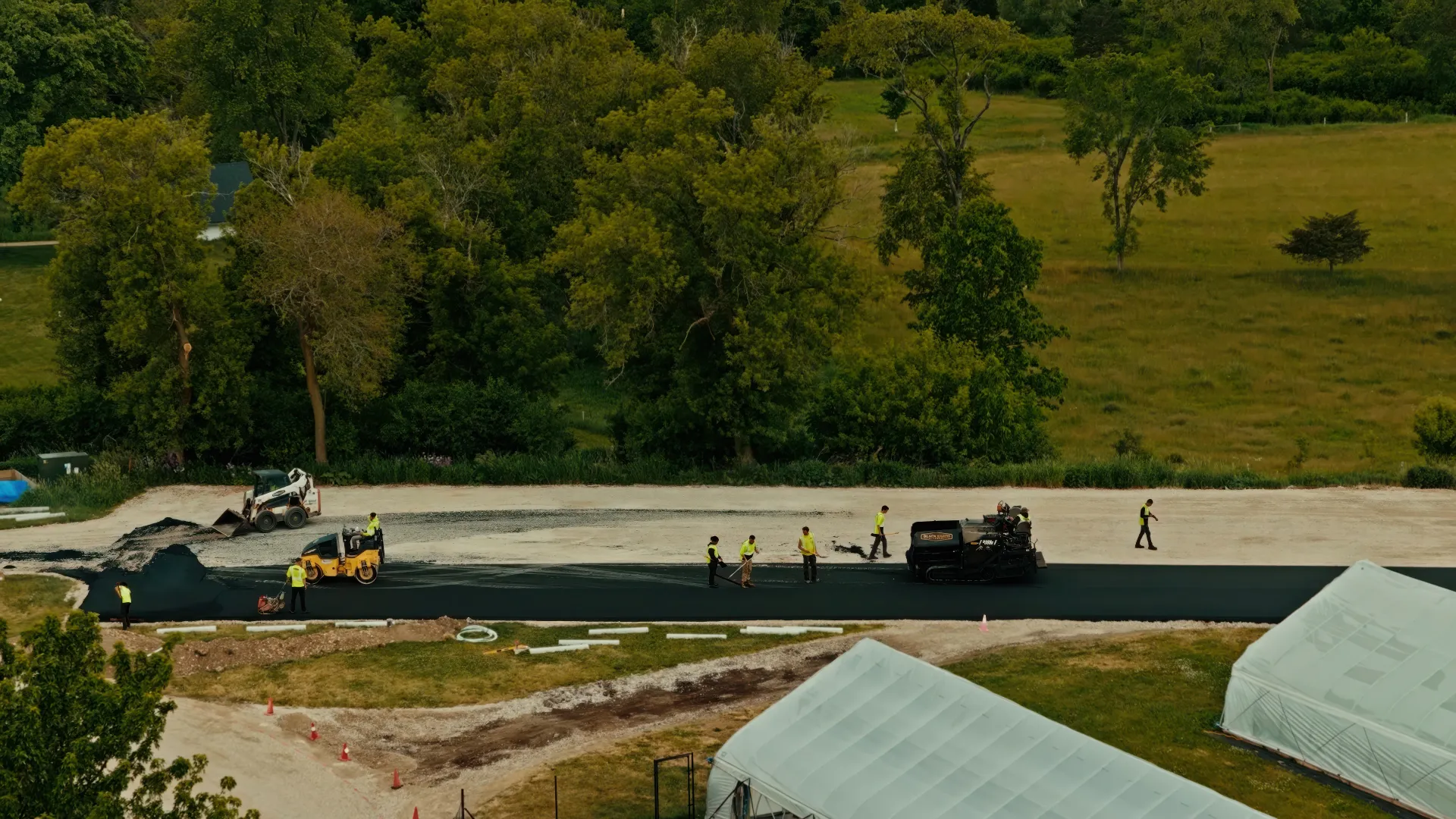 Two construction workers operating compact rollers on freshly paved asphalt near an industrial building.