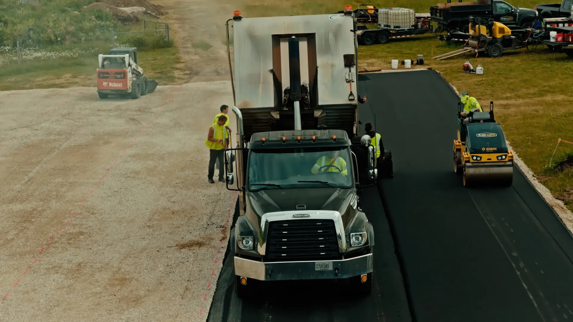 Two construction workers operating compact rollers on freshly paved asphalt near an industrial building.