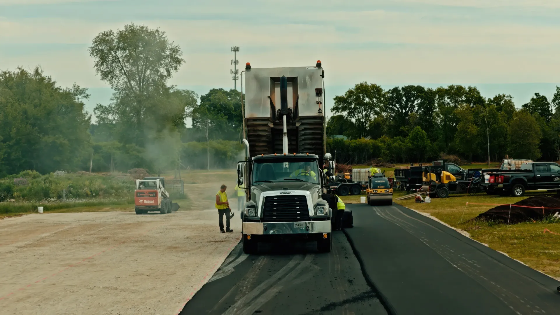 Road paving crew with a large asphalt machine on a rural road, workers nearby and equipment in the background