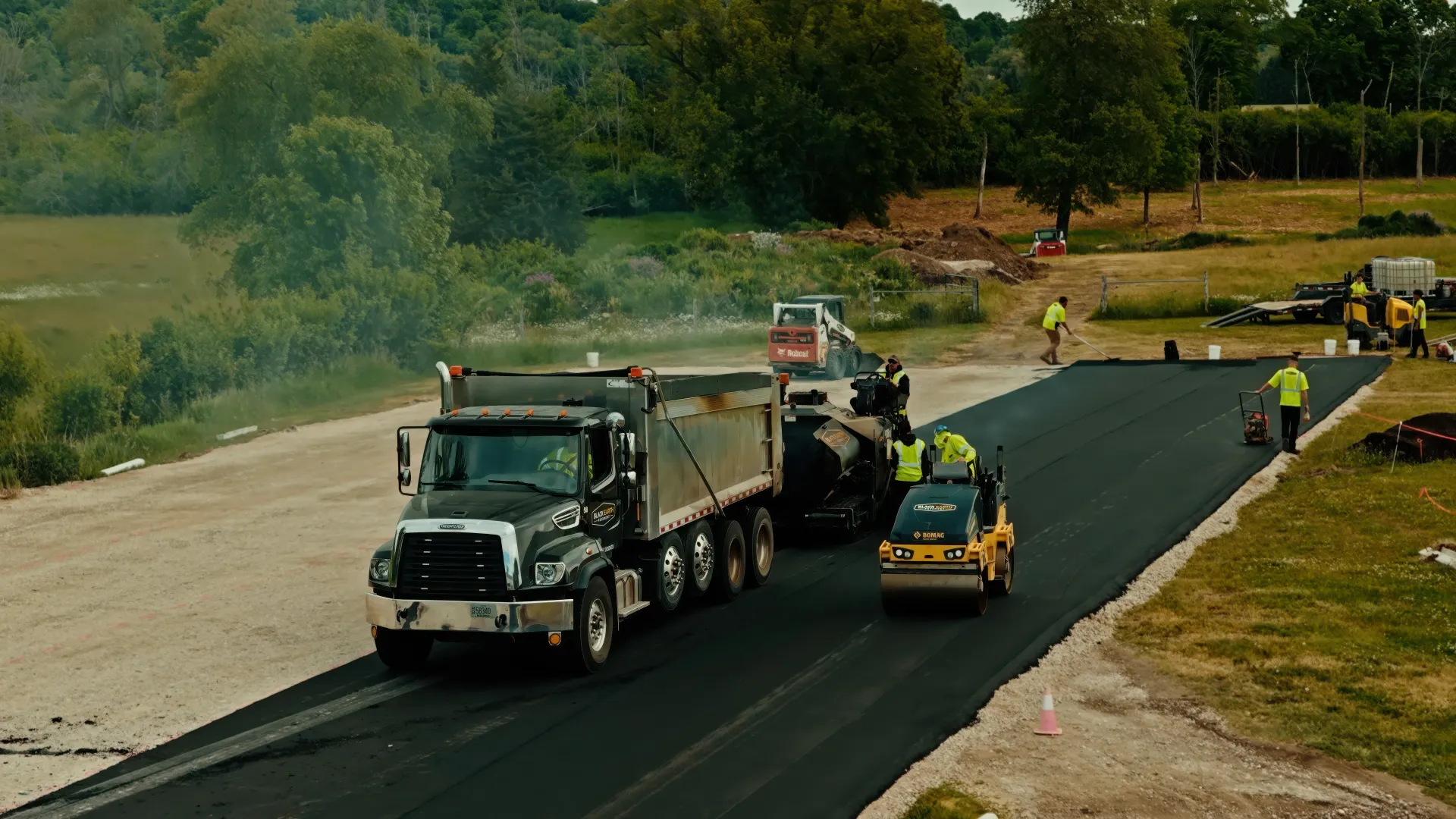 Construction trucks paving a new asphalt road beside a grassy hillside