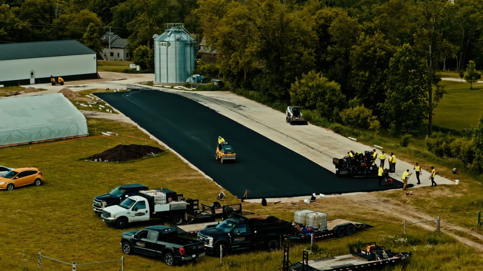 Aerial view of a rural road resurfacing site with workers, vehicles, and construction equipment.