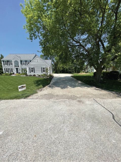 Driveway leading to a white house with a large tree and green lawn on a sunny day