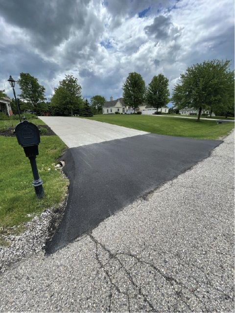 Driveway with fresh asphalt patch leading to a white gravel road under a cloudy sky.