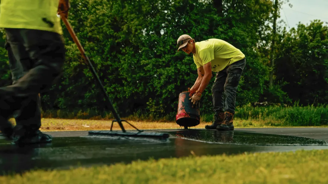 Two workers in neon shirts patching a wet road with a black sealant machine