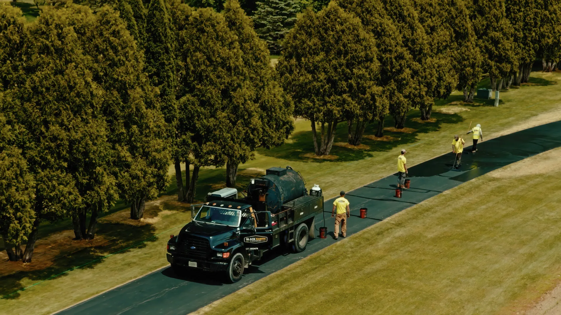 Workers in neon vests surround a truck on a tree-lined roadside, cleaning the shoulder.