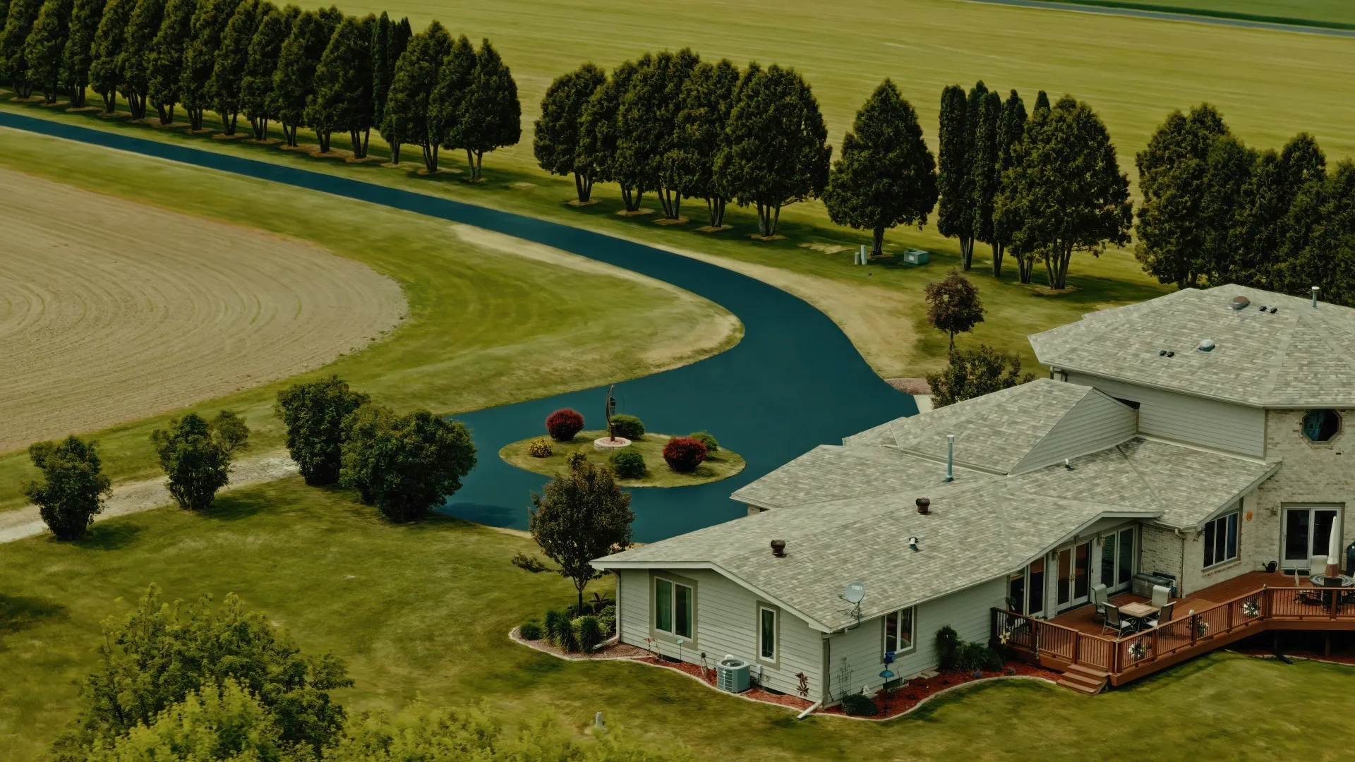 Aerial view of a house beside a winding blue-green stream, with fields and trees around it.
