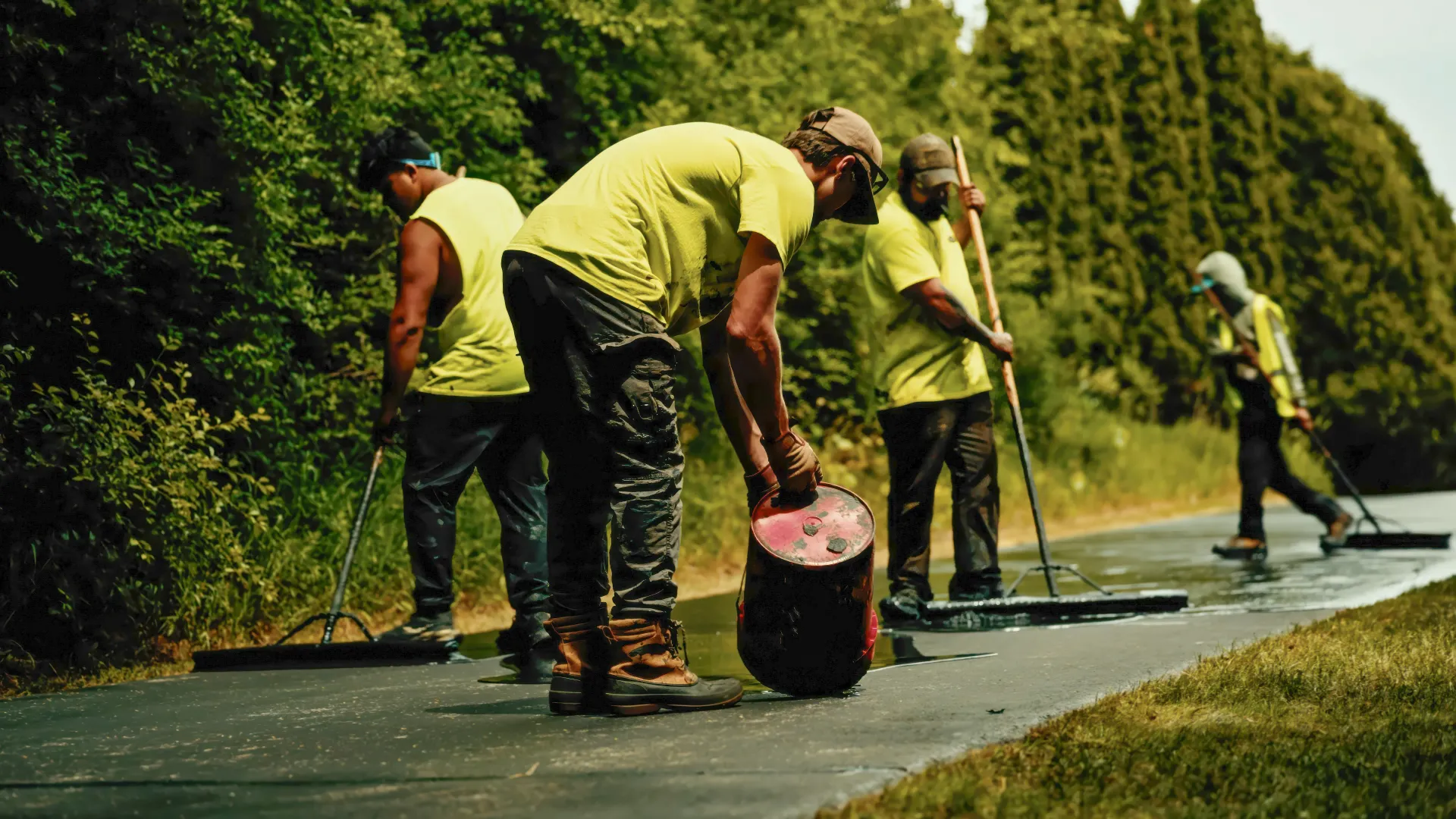 Workers in neon shirts paving a sidewalk beside tall green hedges.