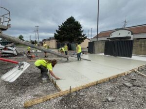 Two construction workers operating compact rollers on freshly paved asphalt near an industrial building.