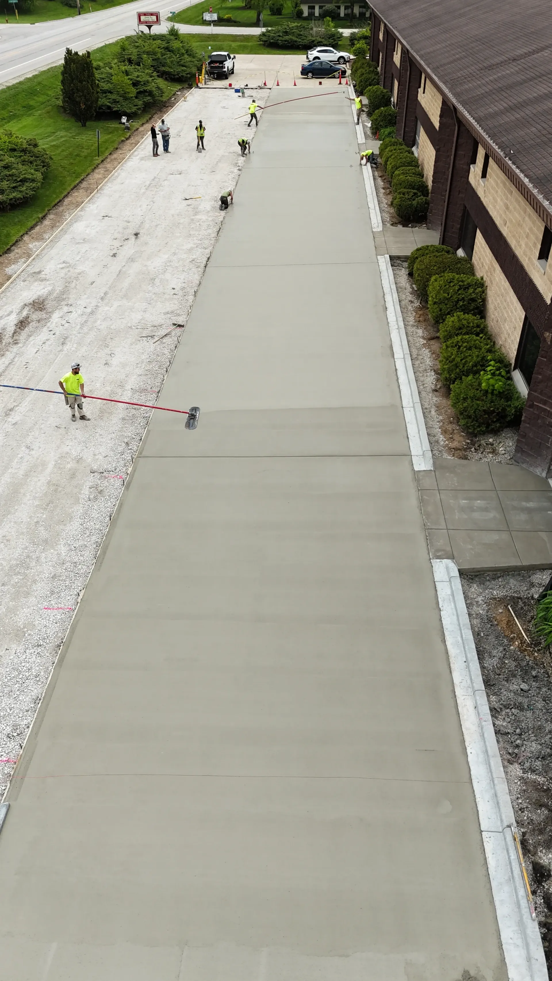 Freshly poured concrete driveway beside a building, with workers and tools visible in the distance.